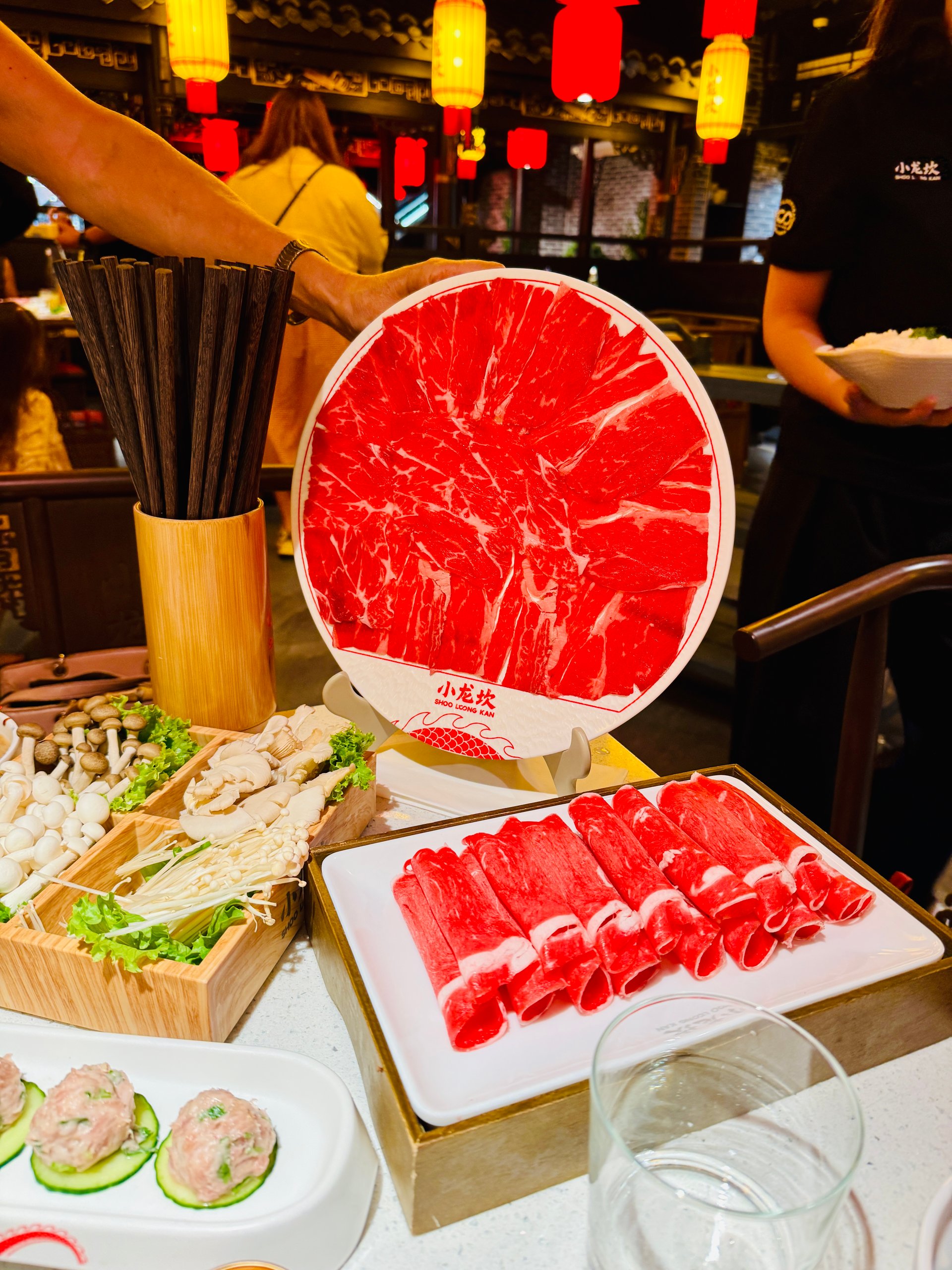 Thinly sliced beef arranged in a circular pattern on a plate, with mushrooms and greens nearby in a restaurant setting.