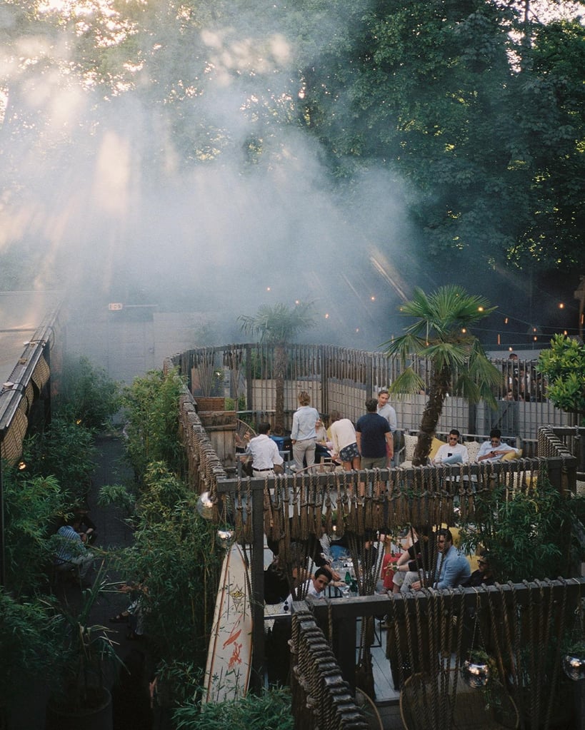 Outdoor rooftop restaurant with wooden railings, string lights, and guests seated at tables amid plants and palm trees, smoke in the air