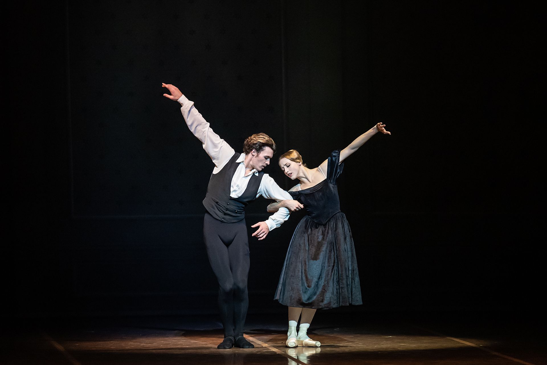 Two ballet dancers perform a duet on a dark stage, the man in a white shirt and vest and the woman in a dark dress with pointe shoes, arms raised in graceful poses.