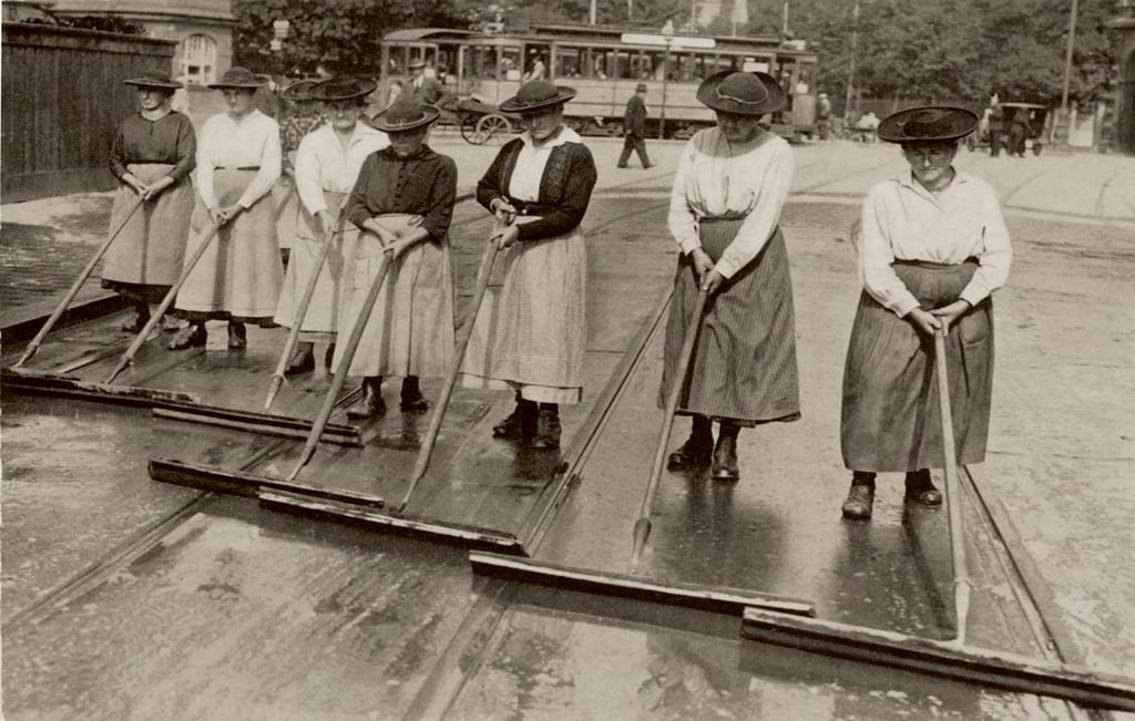 Group of women in early 20th‑century dresses sweeping a wet street with long-handled squeegees; a trolley/tram is in the background.