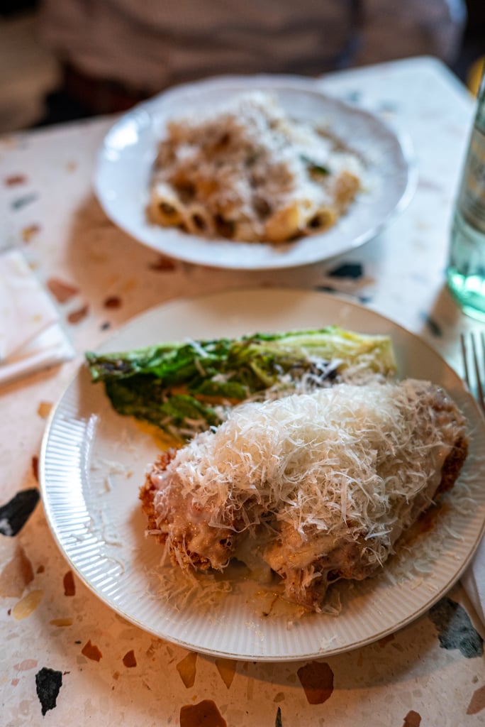 Coconut-crusted fried dish on a white plate seen up close, with a second plate of greens in the foreground on a speckled table.