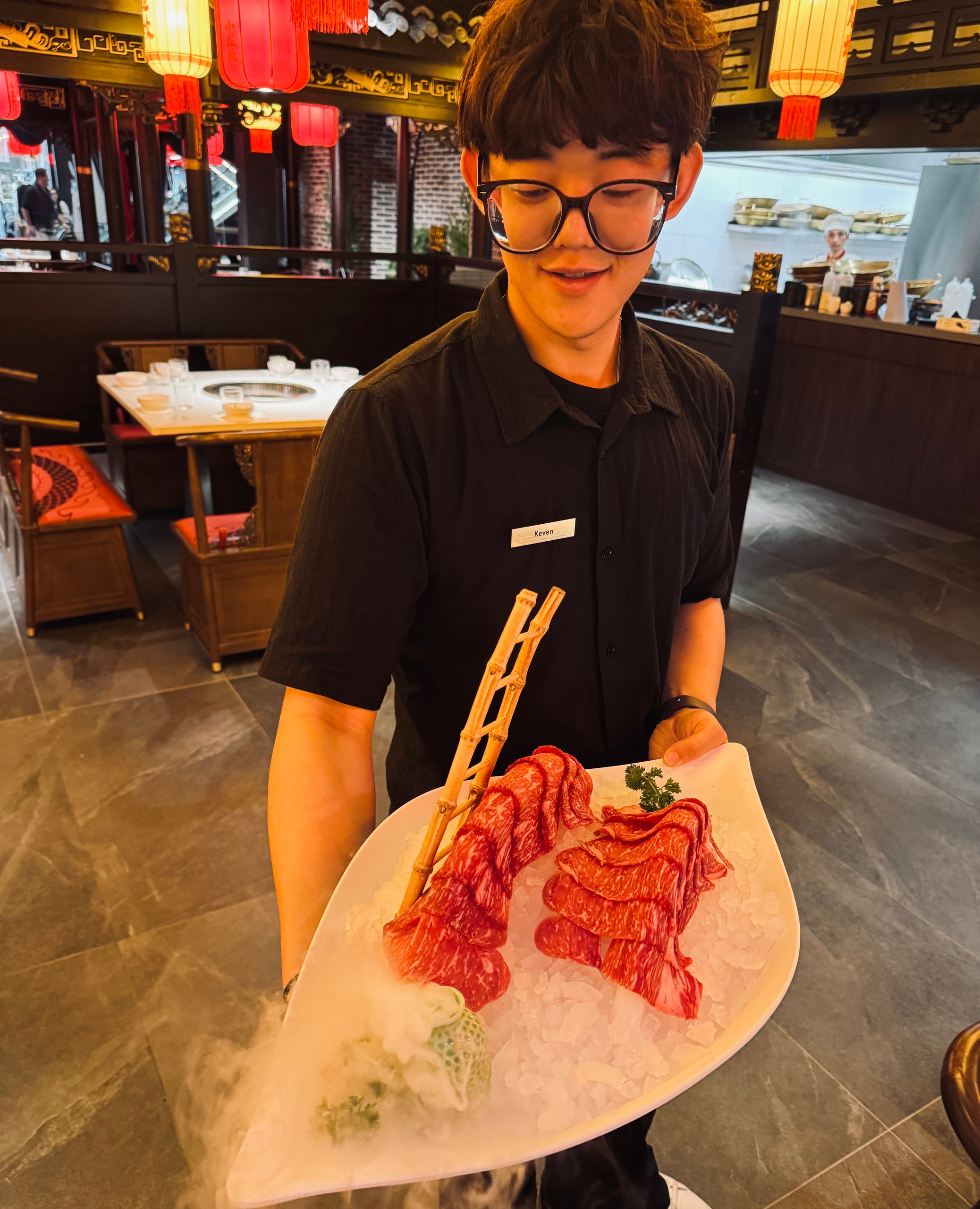 Waiter with glasses presents a plate of thinly sliced beef on ice in a restaurant with red lanterns nearby.