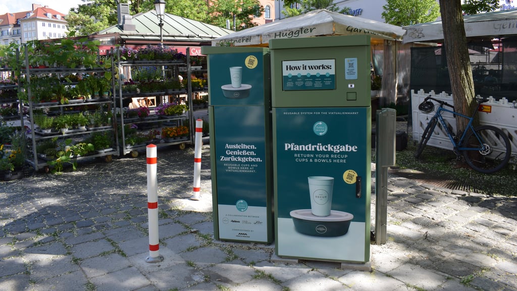 Flower market stalls beside a large green cup return machine labeled 'How it works' in a sunny town square; a blue bicycle rests nearby on the right.