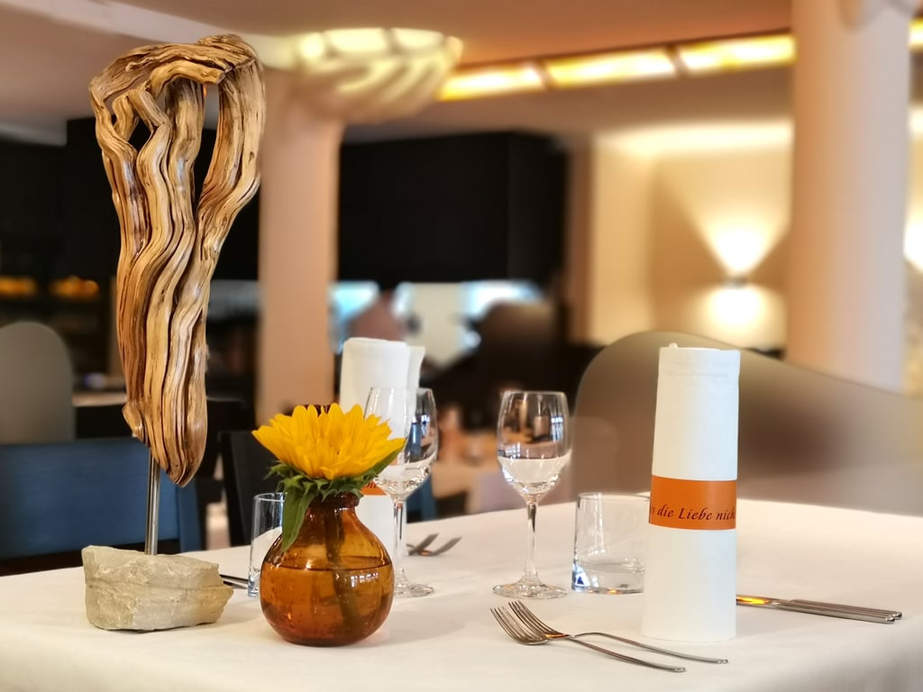Table set for dining with a tall twisted driftwood centerpiece, amber vase with a sunflower, and rolled menus on a white tablecloth, in a softly lit restaurant backdrop