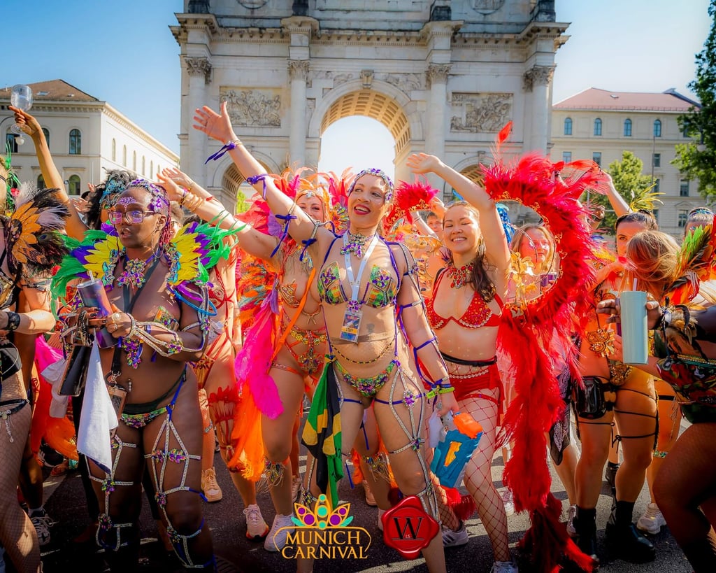 Dancers in bright feather costumes celebrate in a Munich Carnival parade, arms raised with a historic arch behind them.