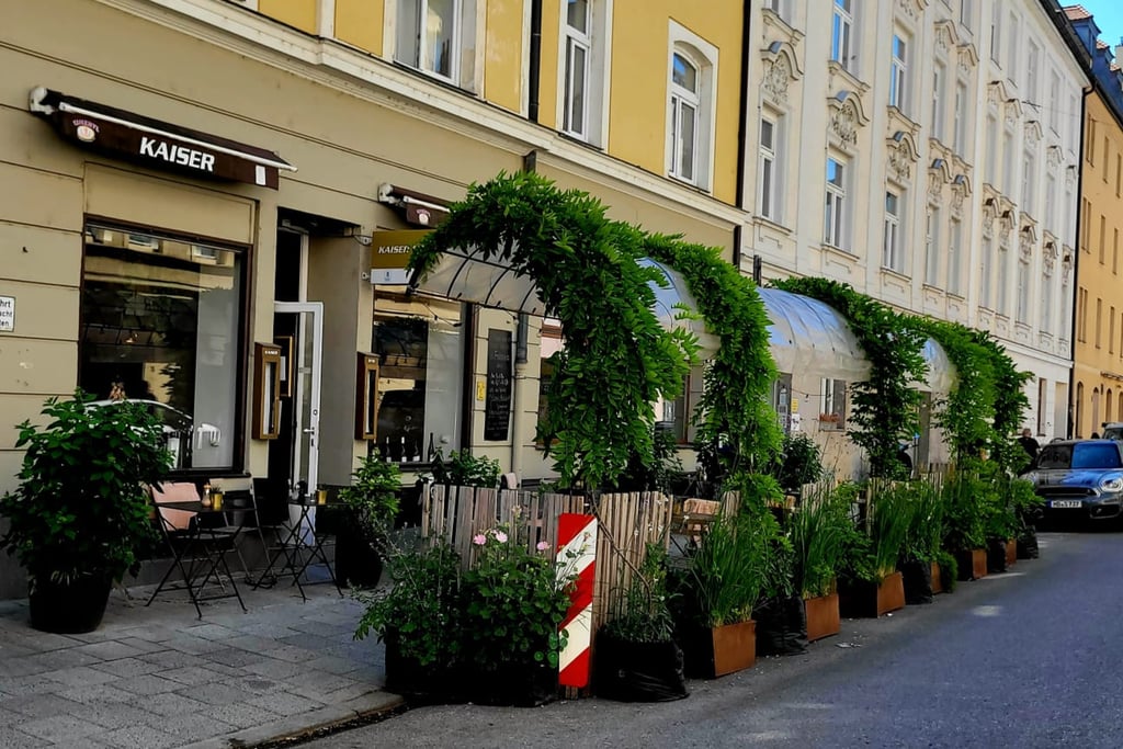 Cafe patio on a narrow street with green leafy arches and potted plants. "Outdoor seating beside a beige building, with green archways and planters."