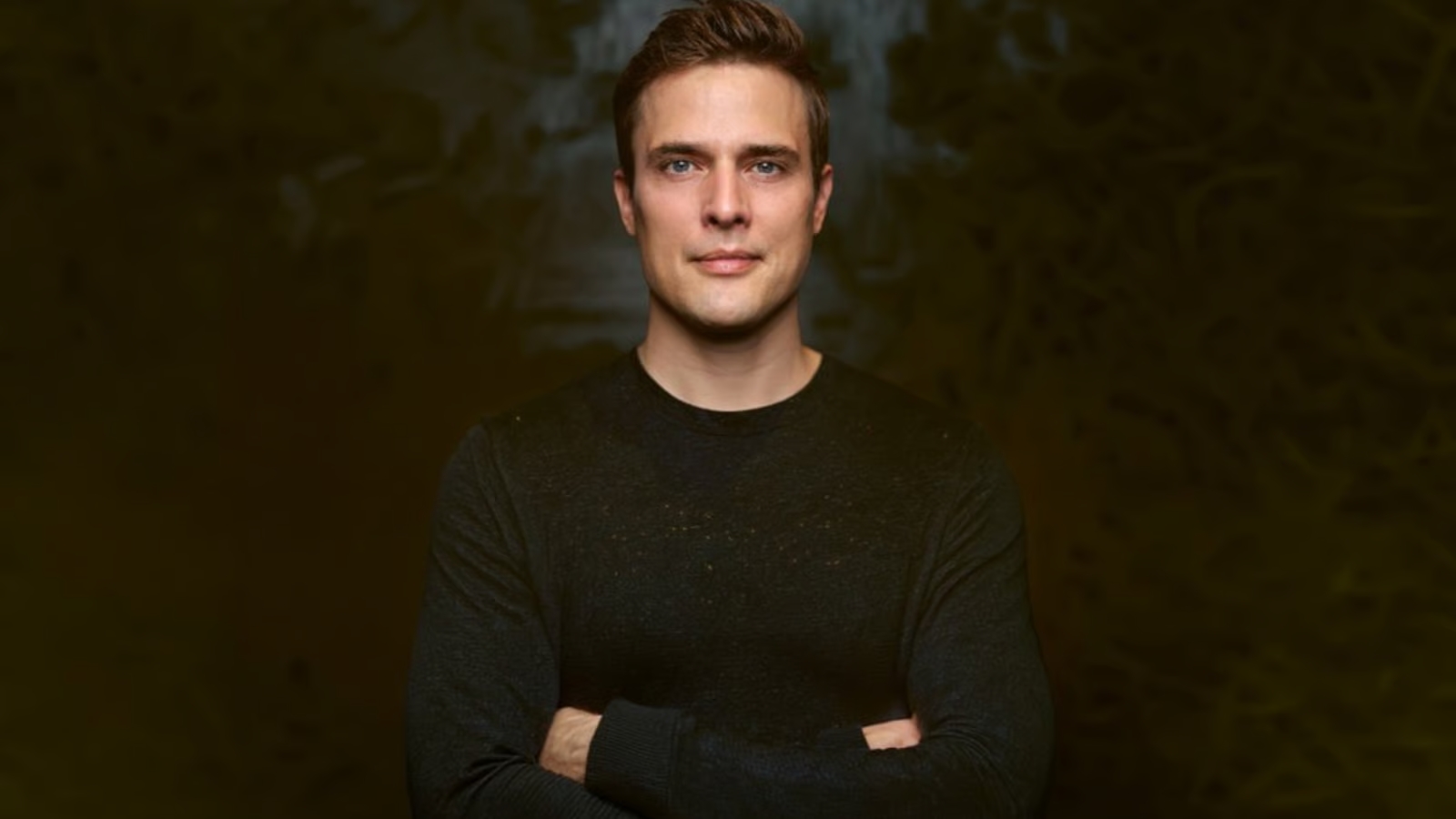 Portrait of a man with short brown hair wearing a black sweater, arms crossed, against a dark blurred background.