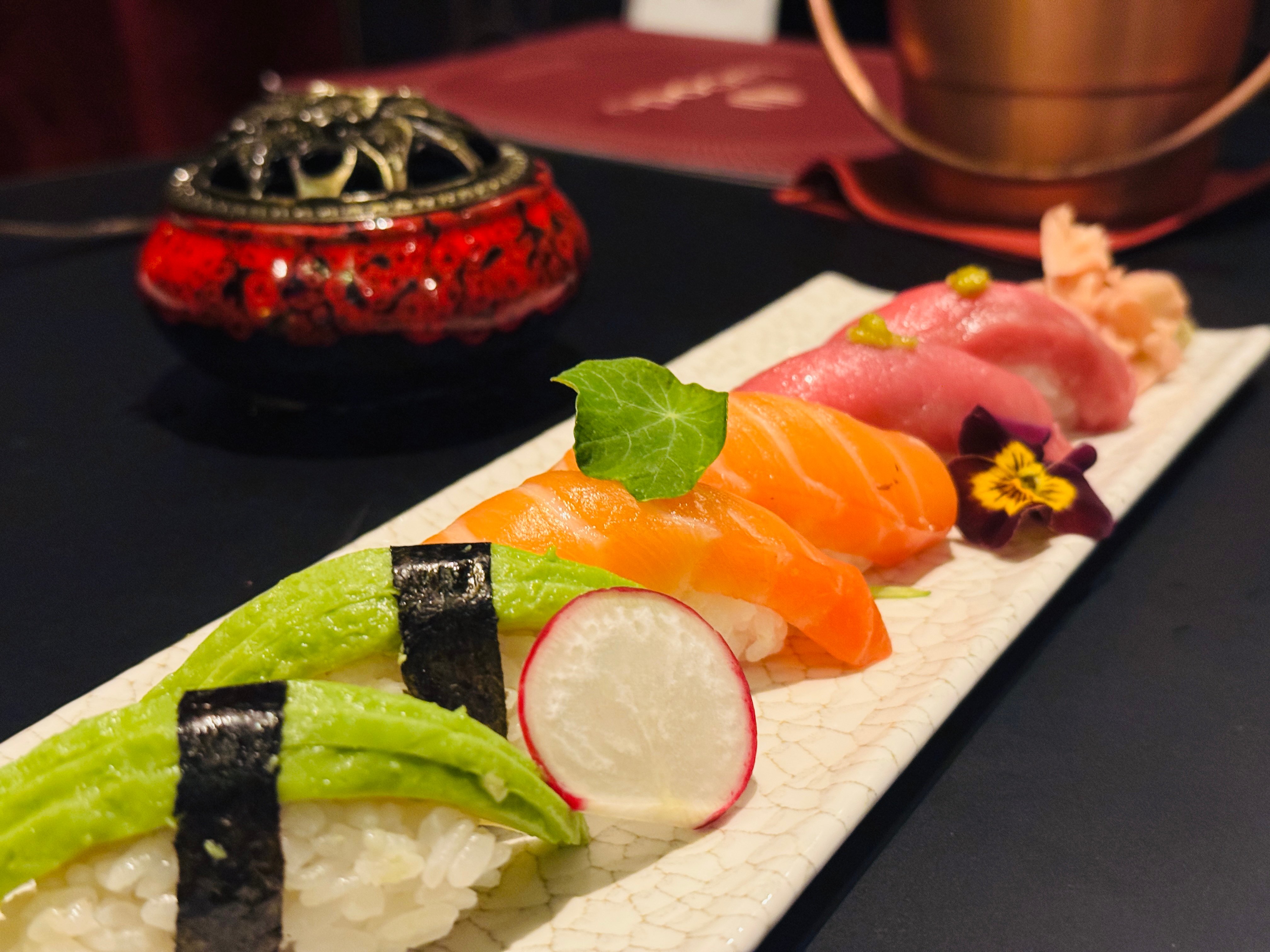 Assorted sushi on a white plate: salmon nigiri, avocado rolls with nori, radish slices, edible flowers; red lacquered box in background.