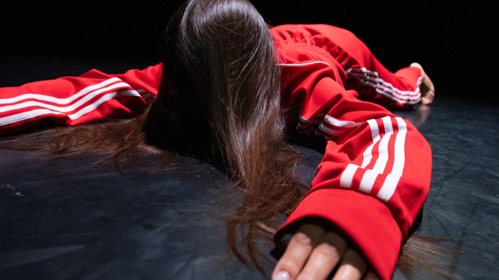 Person in a red track suit with white stripes lies face-down on a dark floor, hair obscuring their face.