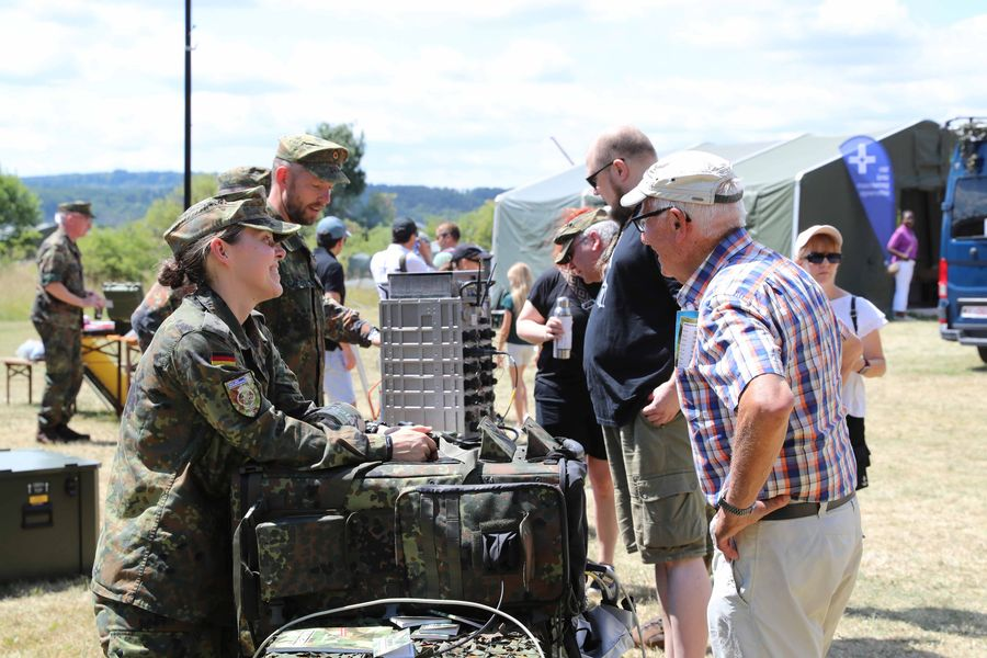 Female soldier in camouflage explains military gear to a line of civilians at an outdoor demonstration, tents in the background.