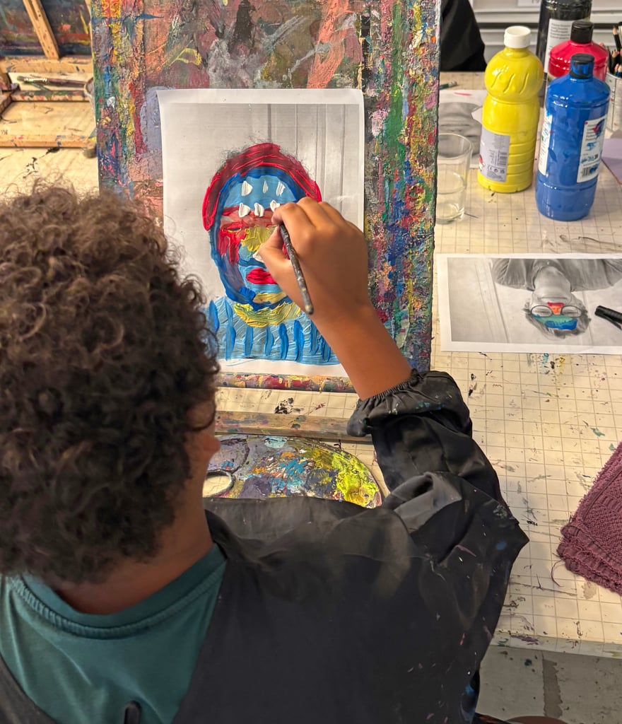 Person painting a colorful portrait on a board, focusing on the brushstroke with bright reds, blues, and yellows; art supplies and paint bottles in a messy studio in the background.