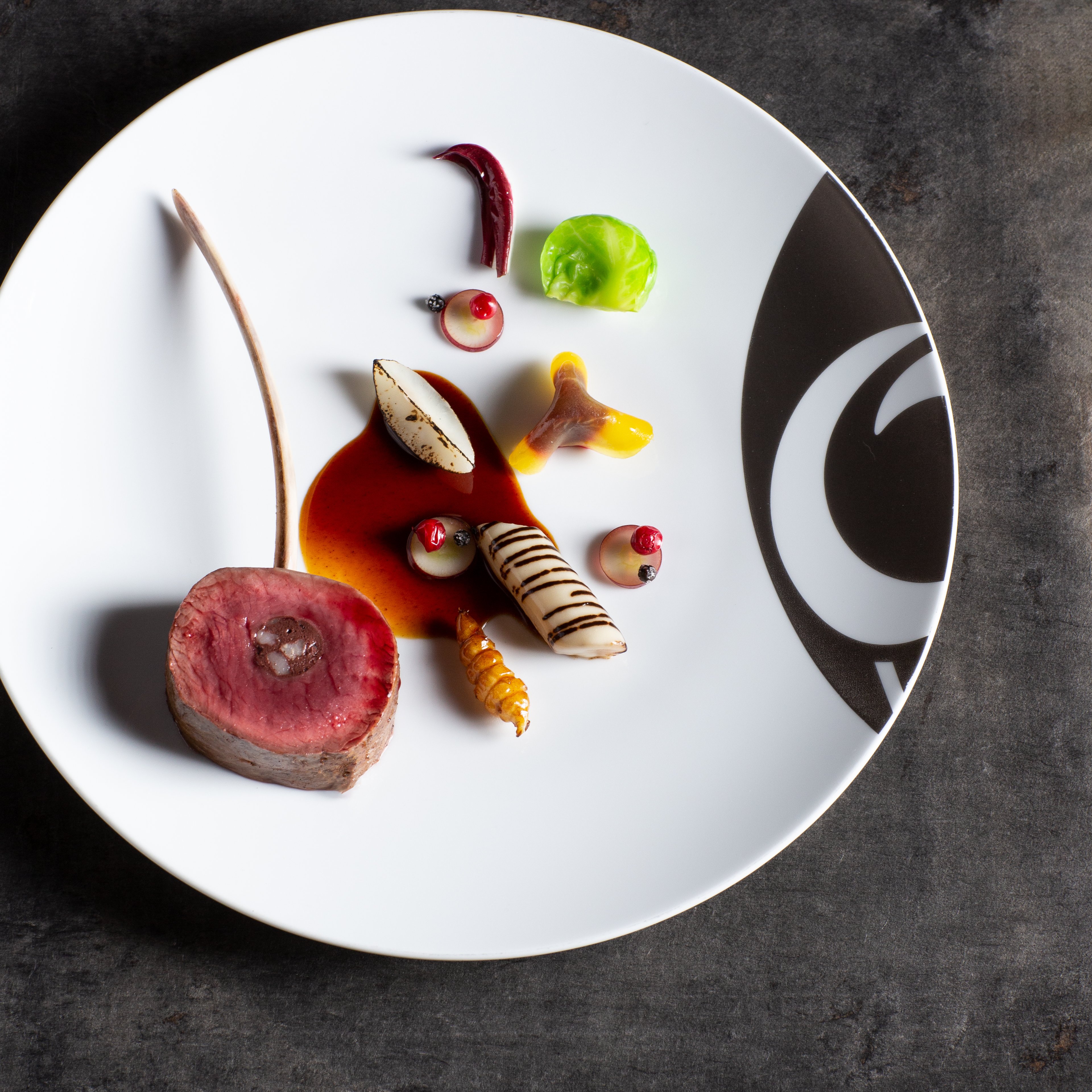Rehfleisch mit Gemüse und Jus auf weißem Teller mit großem Logo am Rand