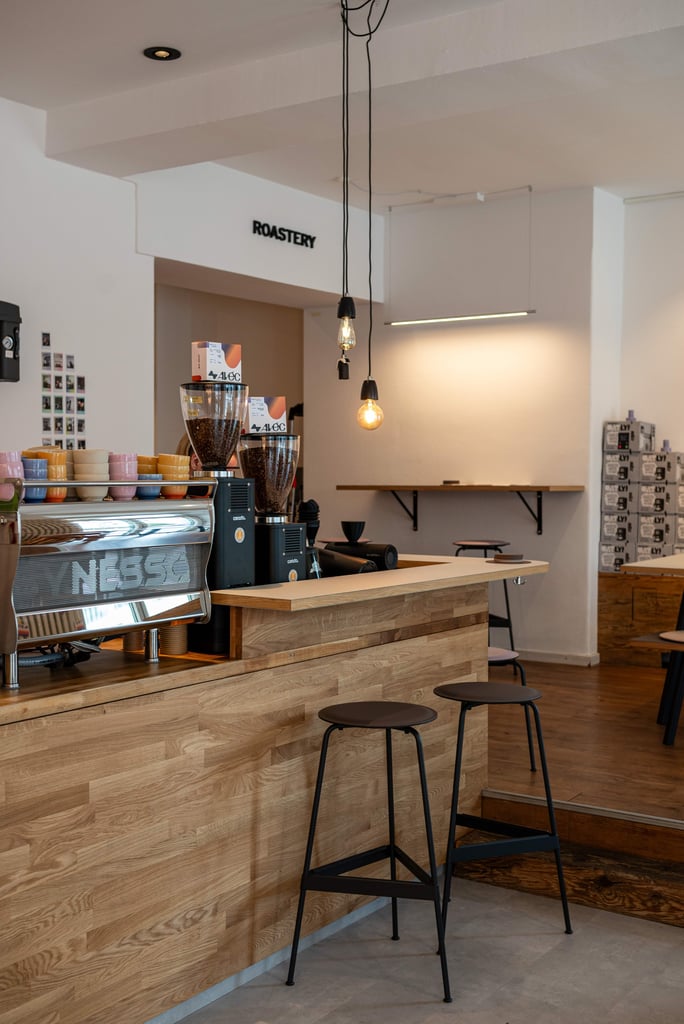 Cafe counter with espresso machine, grinders, and stacked cups; ROASTERY sign on the wall behind. 🡒 Informative and concise description of the setup and branding.