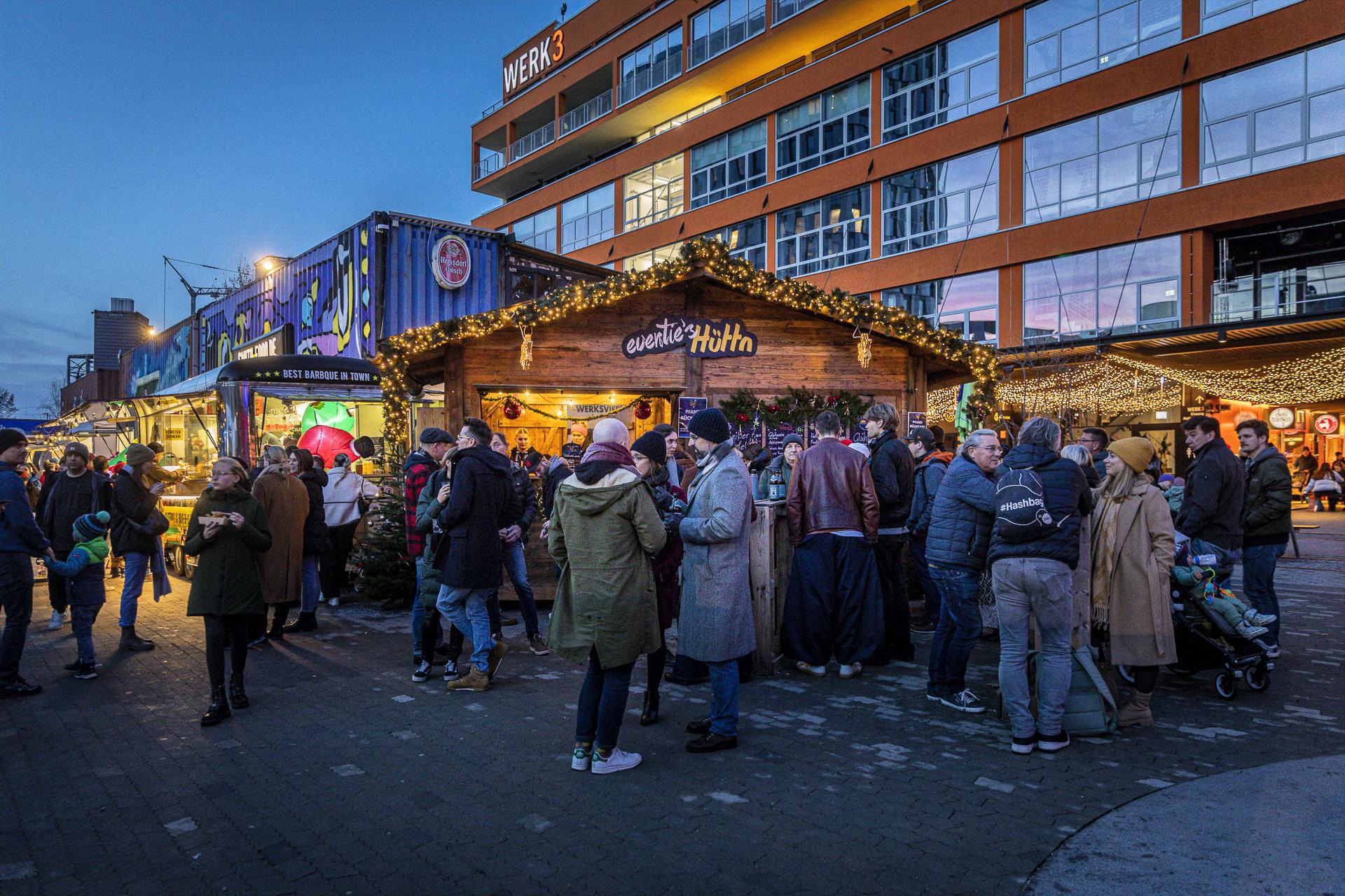 Evening Christmas market with a festively lit wooden stall and a crowd in front of a modern orange building with 'WERK3' signage