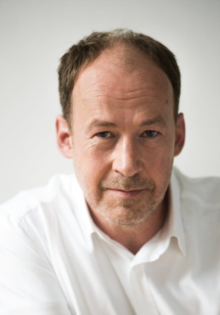 Close-up of a middle-aged man with short brown hair and light stubble, wearing a white shirt against a pale background.