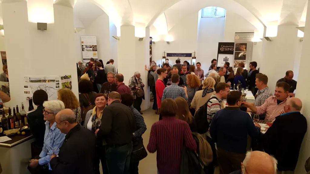 Crowded reception in a bright, arched hall with people mingling around standing tables and booths displaying banners and posters, some sampling wine.
