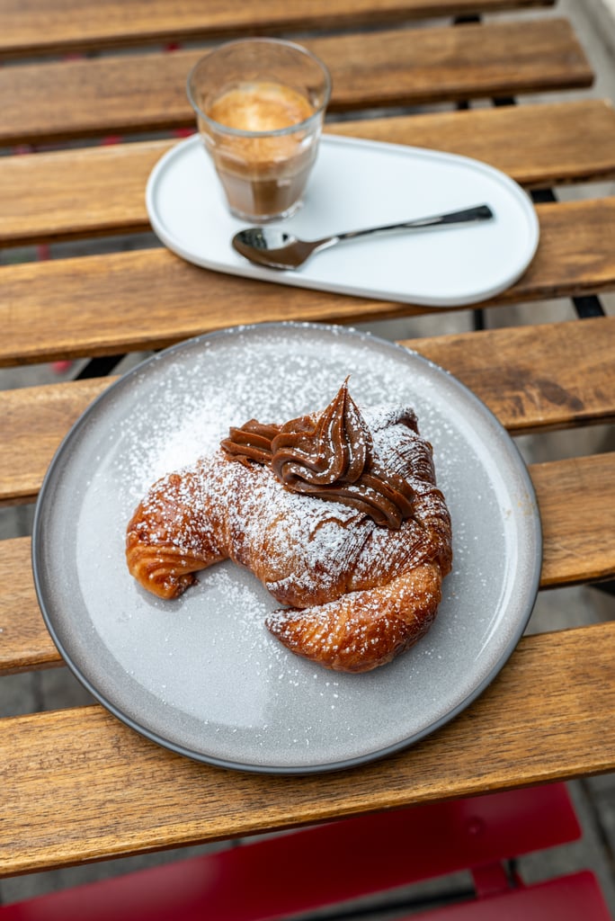 Powdered-sugar-topped chocolate croissant on a gray plate with a cup of coffee in the background on a wooden table on a sunny day.