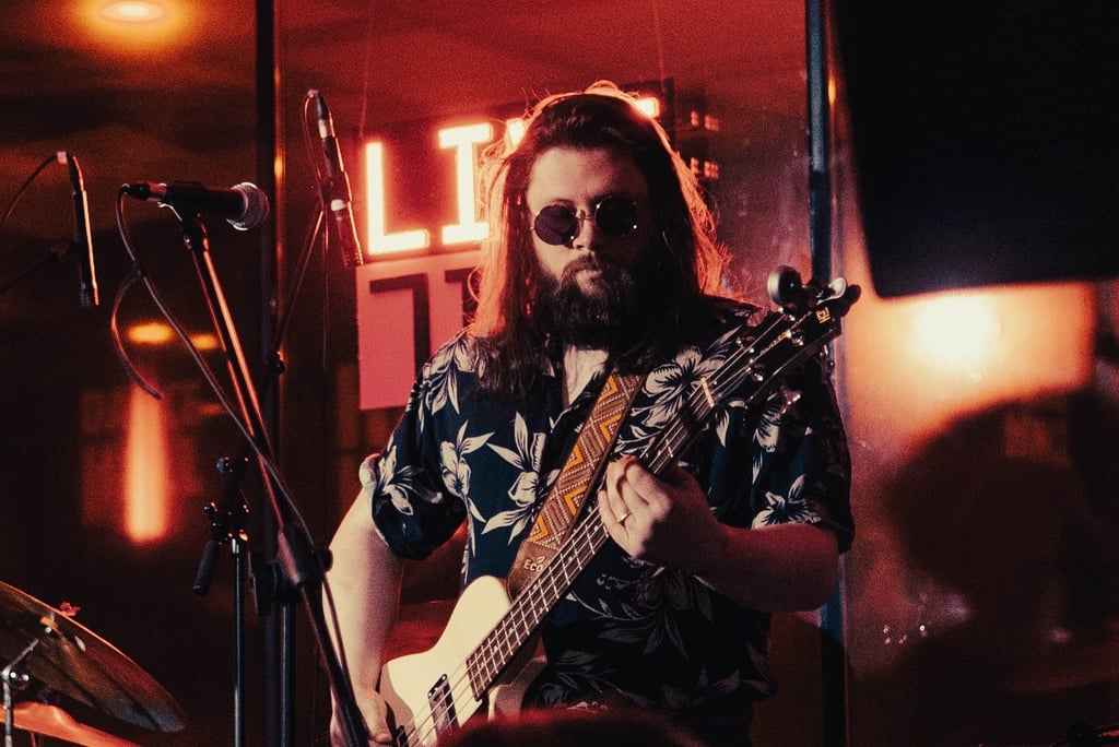 Musician with long hair and beard wearing sunglasses, playing a bass on a dim stage with red neon light behind.
