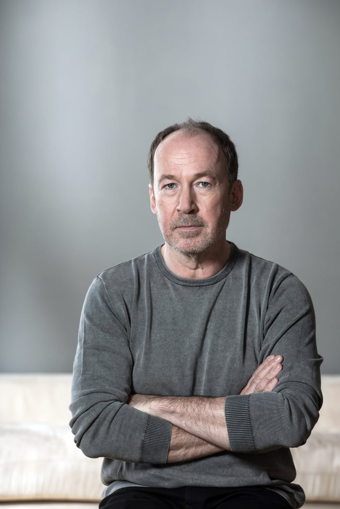 Portrait of a middle-aged man with a gray sweater, arms crossed, sitting against a neutral gray background, looking at the camera with a calm expression.