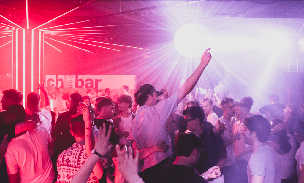 Crowded nightclub with red and purple lighting, people dancing and raising hands under a bright disco ball.