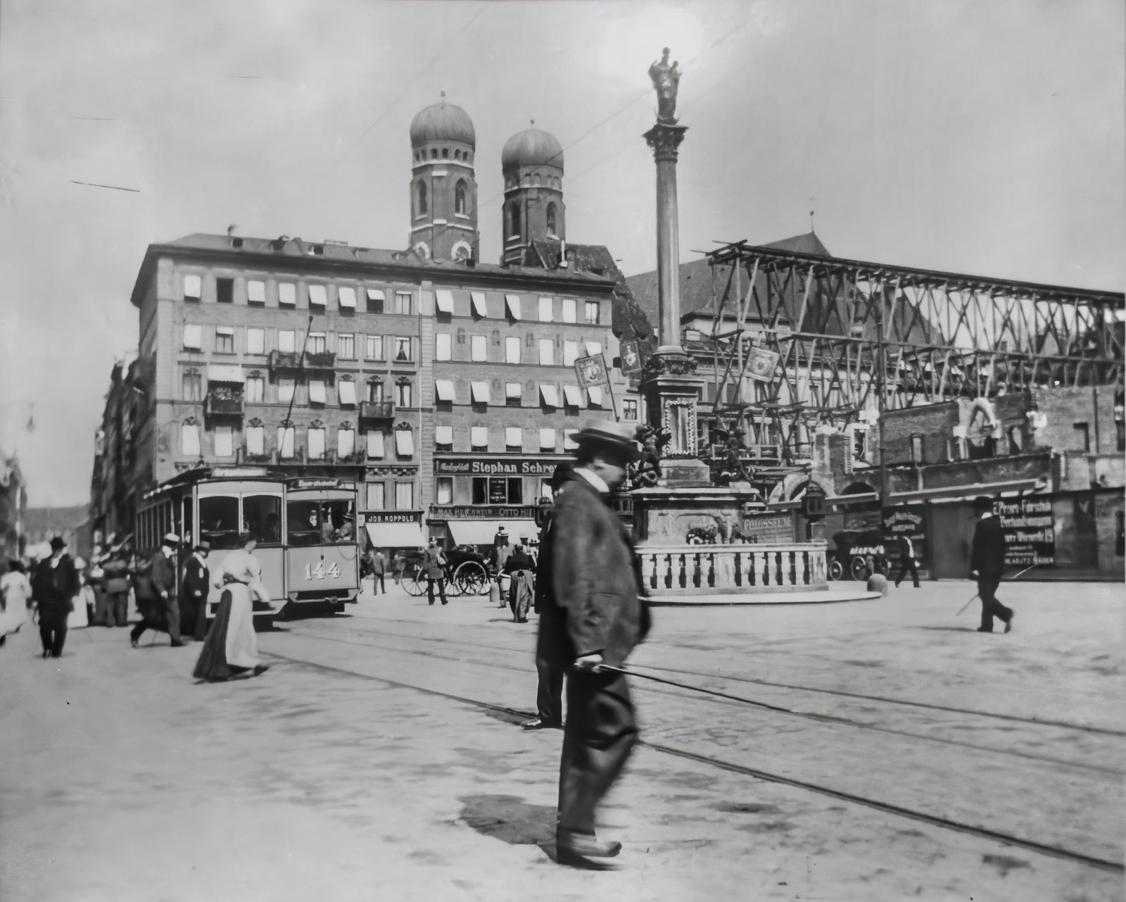 Black-and-white city street scene with tram tracks, brick buildings, and a tall statue column in the center; pedestrians in foreground.