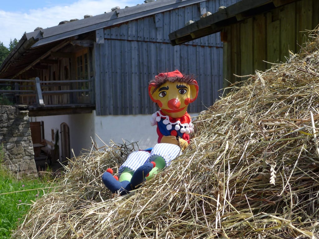 Clown-faced scarecrow puppet with red hat among dry straw, with a wooden barn behind it.