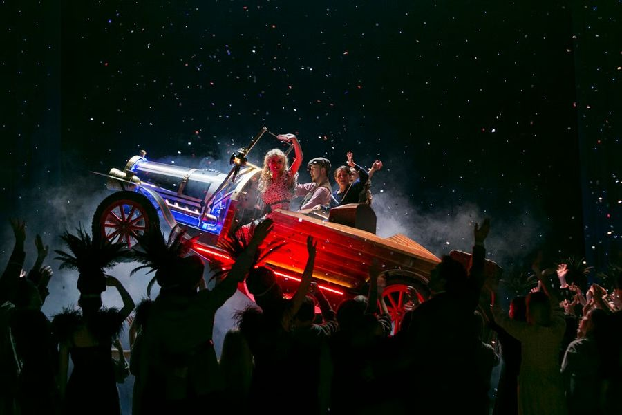 A neon-lit vintage car prop on a stage with performers posing inside as a cheering crowd raises their hands and confetti falls around them.