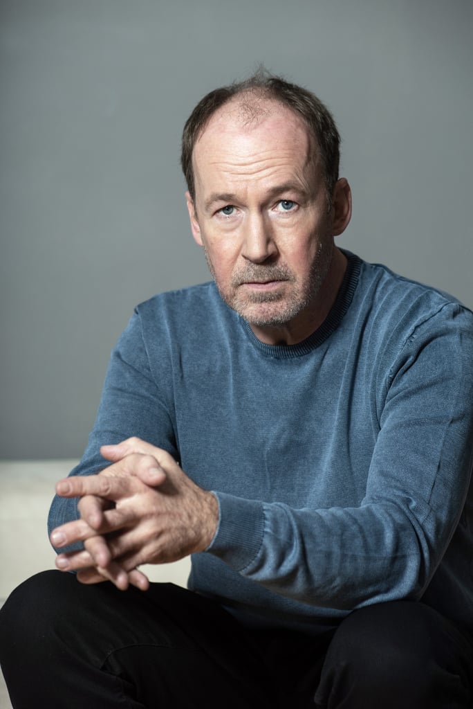 Middle-aged man with light stubble, wearing a blue sweater, seated with hands clasped and a serious expression against a gray background.