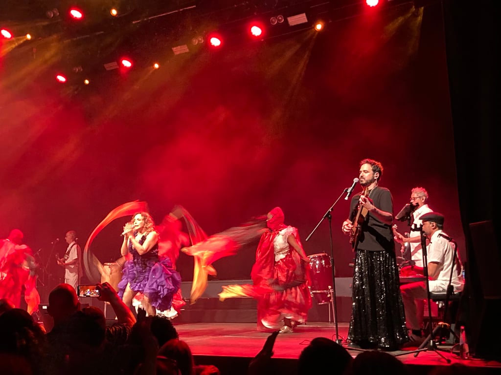 Stage performance with a singer at the microphone, guitarist, and dancers in vibrant costumes under red stage lights. (Informative)