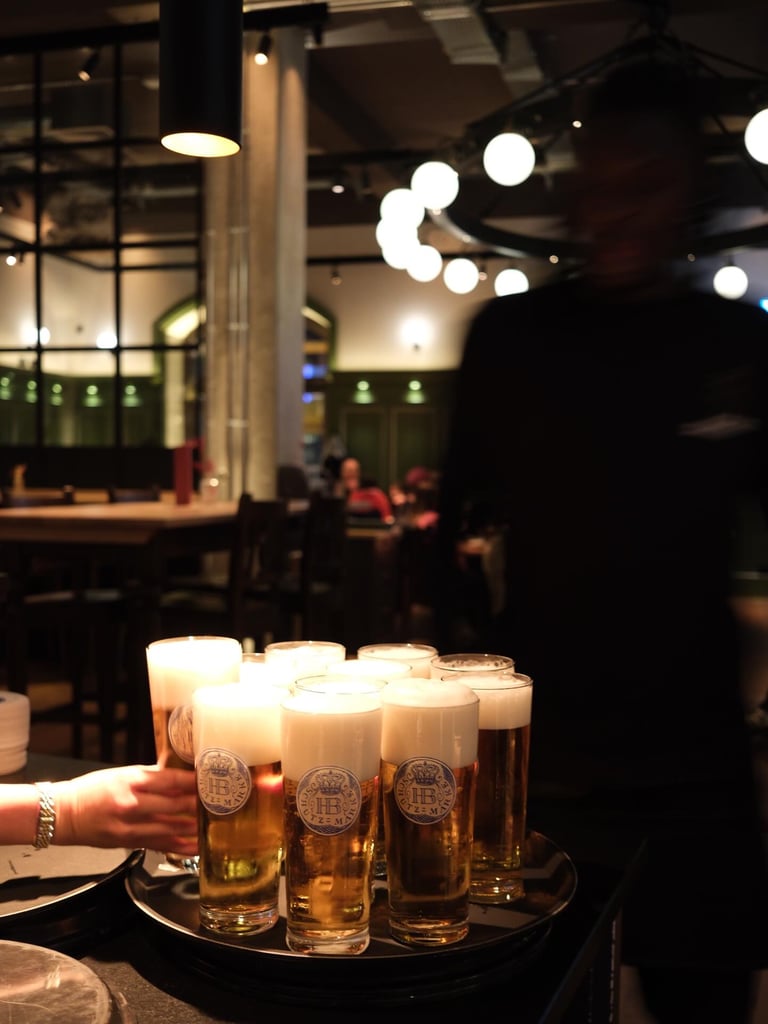 Row of beer glasses with frothy heads on a round tray at a dimly lit bar, warm candlelight in the foreground and blurred patrons in the background.