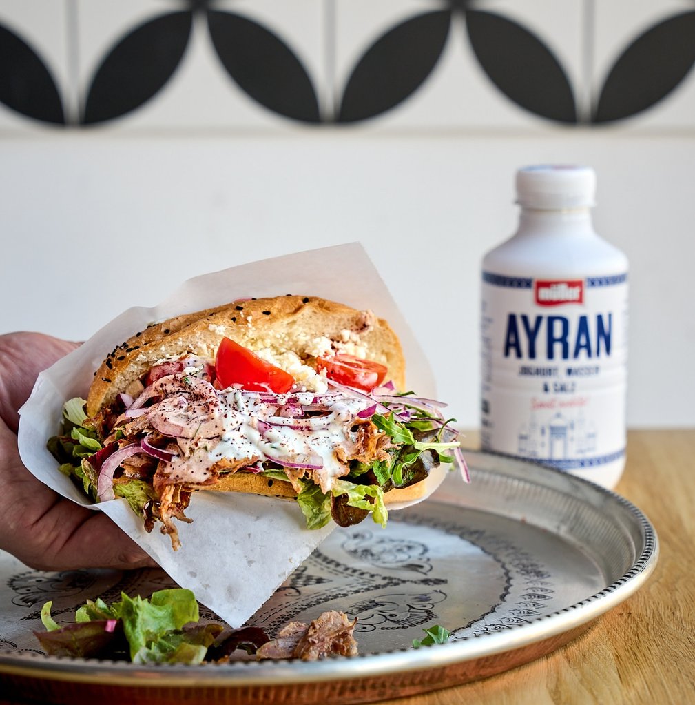 Hand holds a sesame bun sandwich with chicken salad, lettuce, tomato and red onion; Ayran bottle in the background.