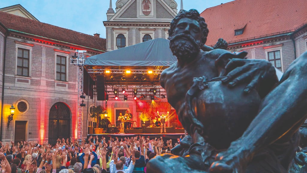 Outdoor concert in a historic plaza with a large statue in the foreground and a band performing on a illuminated stage with cheering crowd watching from below