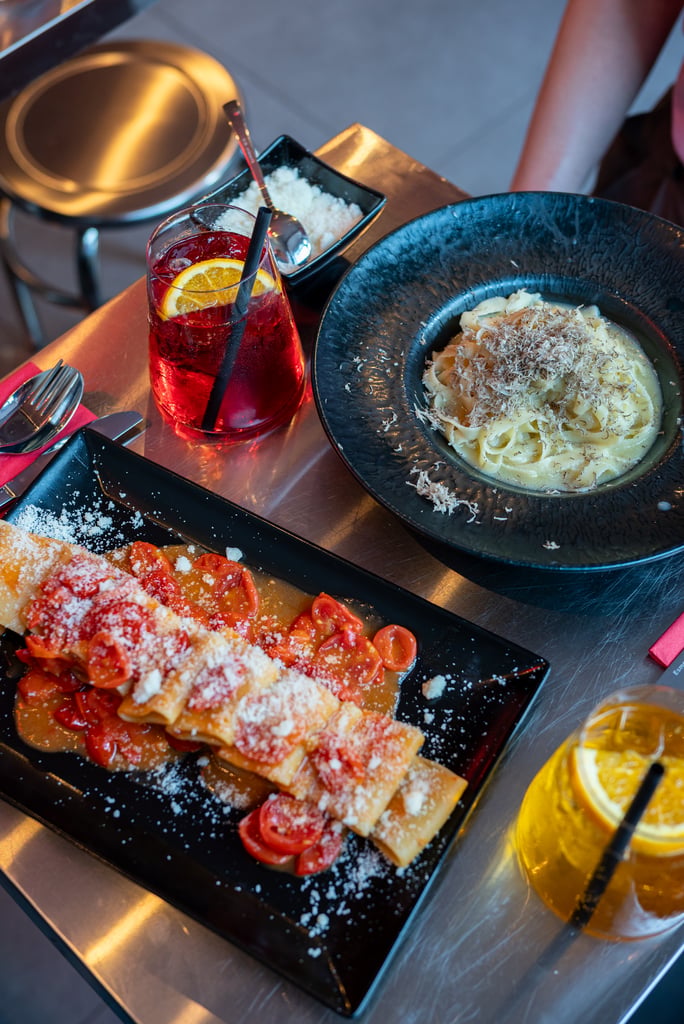 Black plate of pasta with tomato sauce and grated cheese, with a red citrus cocktail beside it on a metal table.