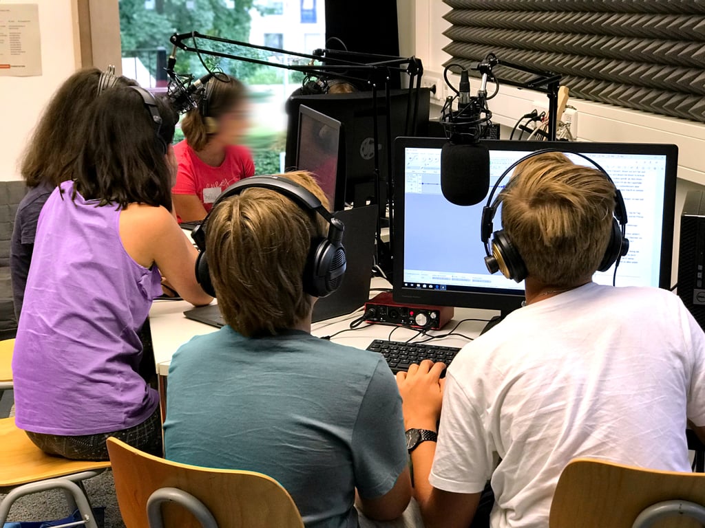 Group of children wearing headphones around desks in a recording studio with microphones and computer monitors nearby.