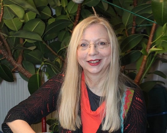 Smiling woman with long blonde hair and glasses, wearing a red top, seated indoors with large plant leaves behind.