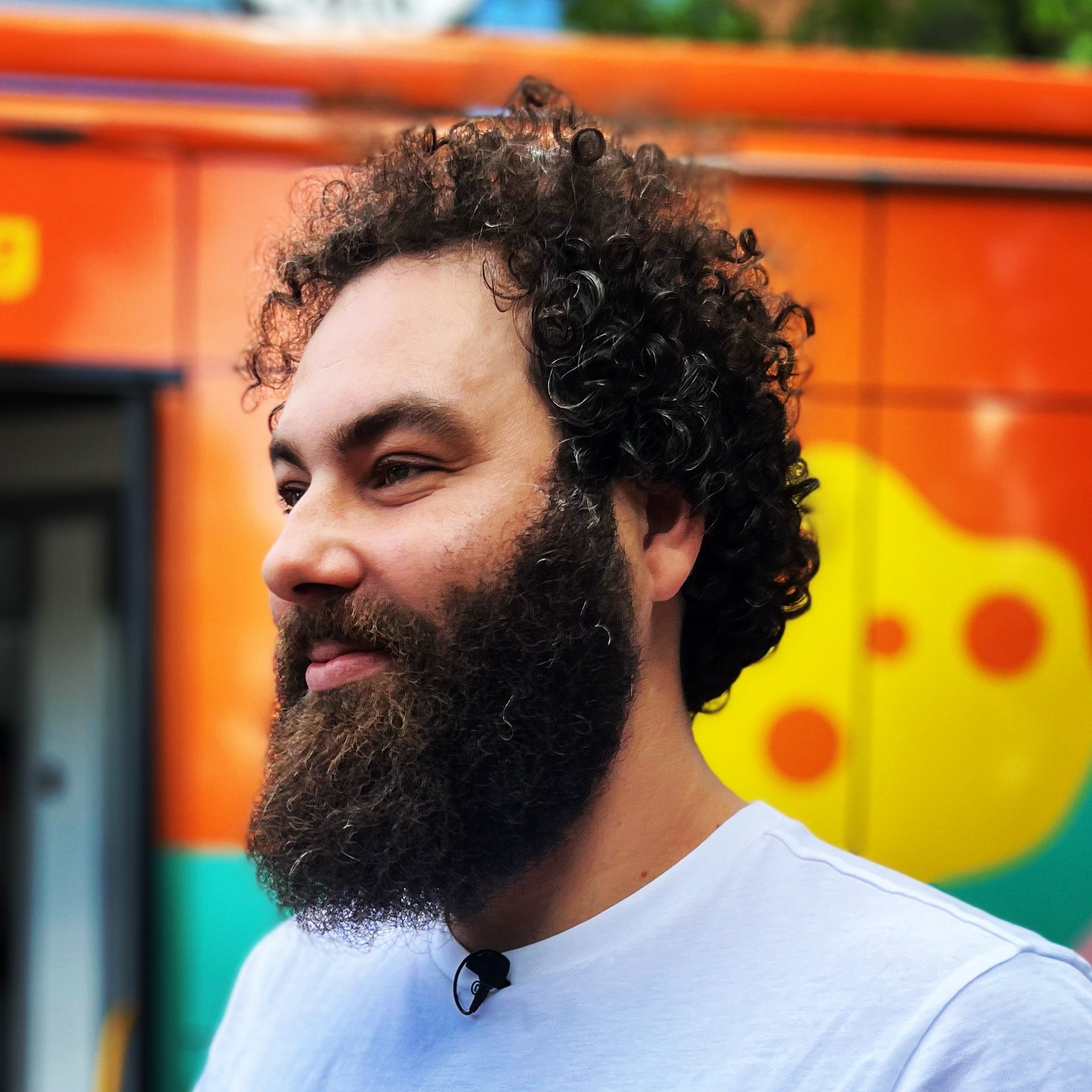 Close-up of a bearded man with curly hair wearing a white shirt and a lapel mic against an orange background.