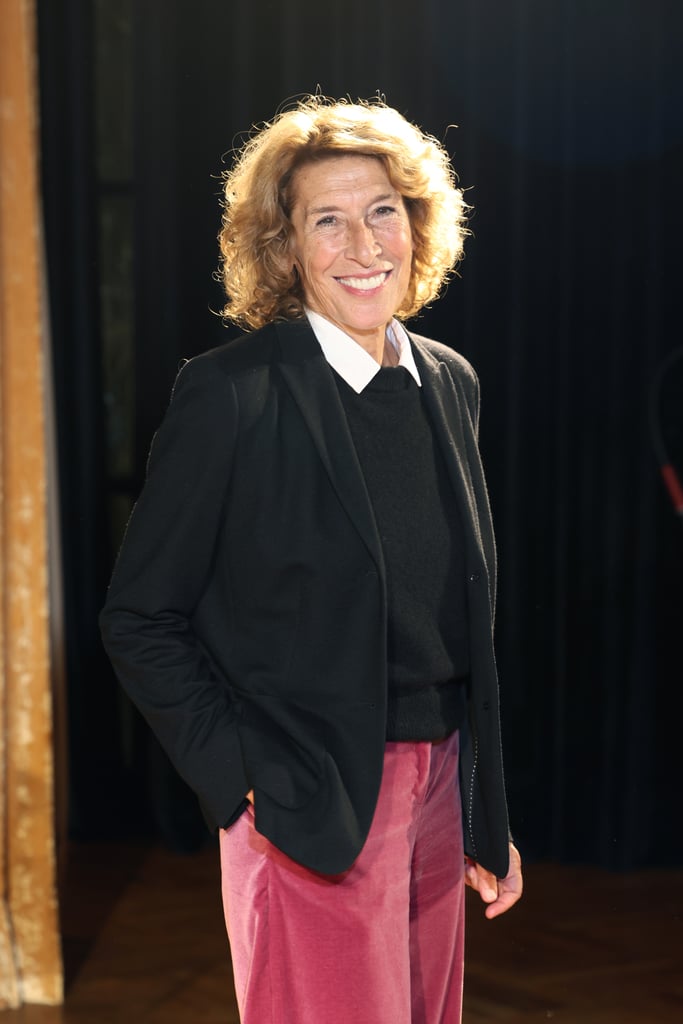 Smiling woman in black blazer and white-collared shirt standing in a doorway, wearing pink pants against a dark background.