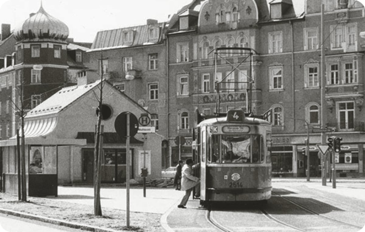 Black-and-white street scene with a vintage tram stopped at a curb, passengers boarding, lined by old brick buildings and a waterfront shopfront.