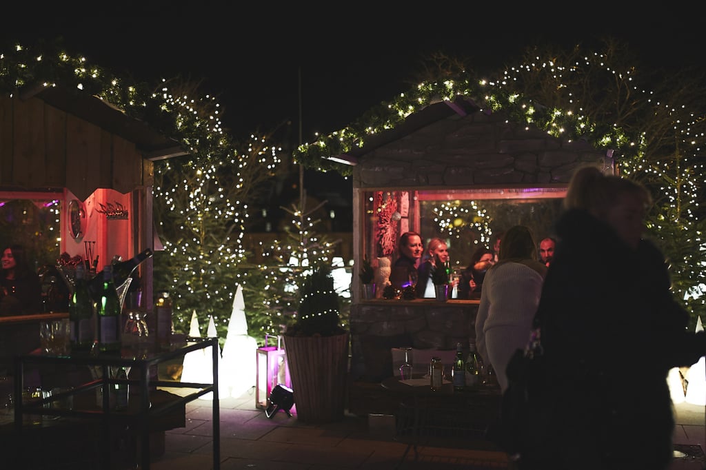 Night scene of a festive outdoor market with twinkling string lights, wooden stalls, and people inside a stone booth chatting.