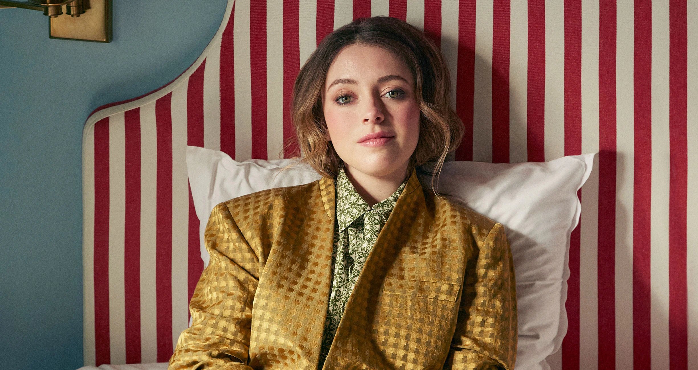 Woman in a gold patterned blazer seated on a bed with white pillows, in front of a red-and-white striped wall and teal background.