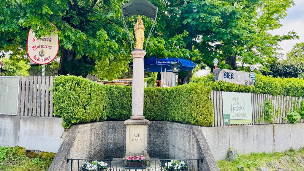 Stone pedestal with 'AVE MARIA' inscription and a statue on top, set in a garden with green hedges and a concrete base nearby.