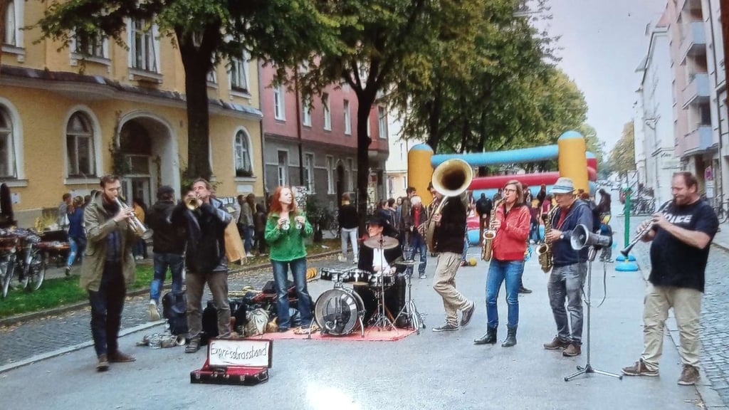 Street musicians play brass and drums on a city street while a small crowd watches, with a colorful inflatable slide in the background and yellow building façades to the left.