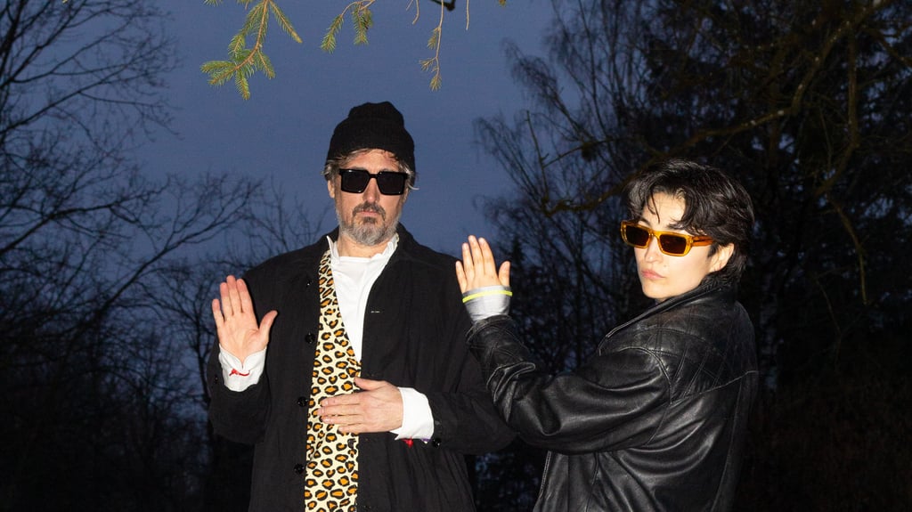 Two adults posing outdoors at dusk; man wearing a beanie, sunglasses, white shirt, leopard-print tie and black coat raises his hands while the woman beside him, in a black leather jacket and orange sunglasses, gestures with her arm.
