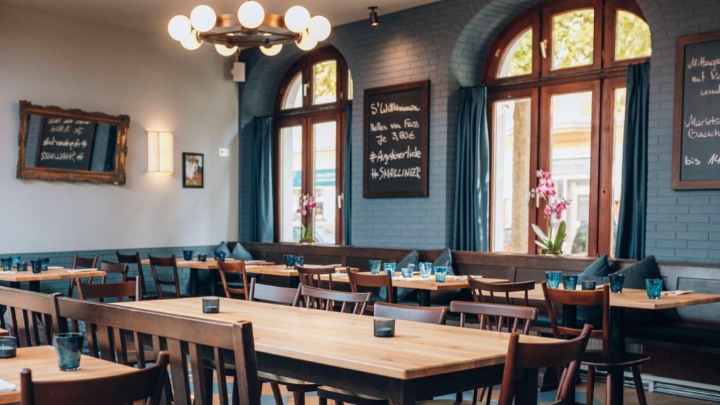 Cozy cafe interior with wooden tables and chairs, chalkboard menus, and blue curtains by arched windows.