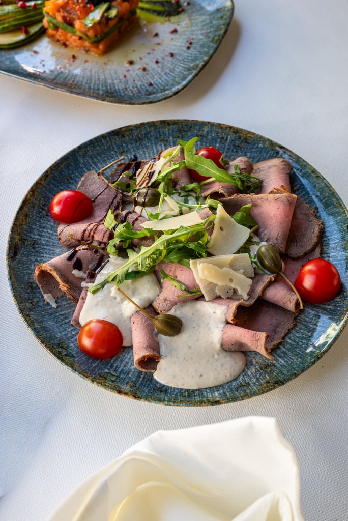 Sliced roast beef with arugula, shaved cheese, cherry tomatoes, and a drizzle of balsamic on a blue plate with creamy sauce nearby
