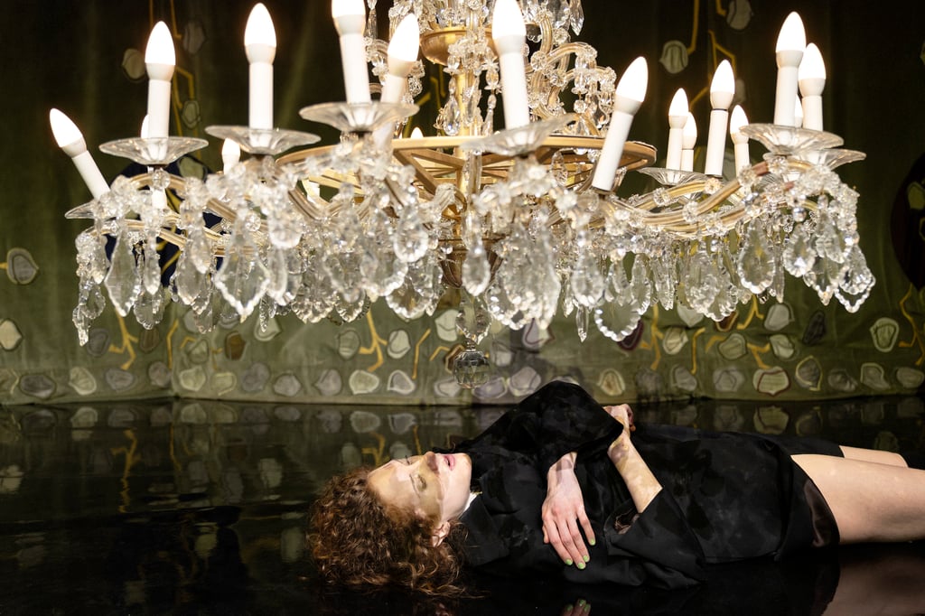 Woman in a black gown lies on a glossy floor beneath a large crystal chandelier, emerald-green nails visible.