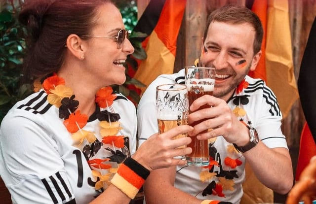 Two friends in sports jerseys toast with beer glasses at a festive outdoor party, wearing orange-black-gold leis and smiles on a sunny day