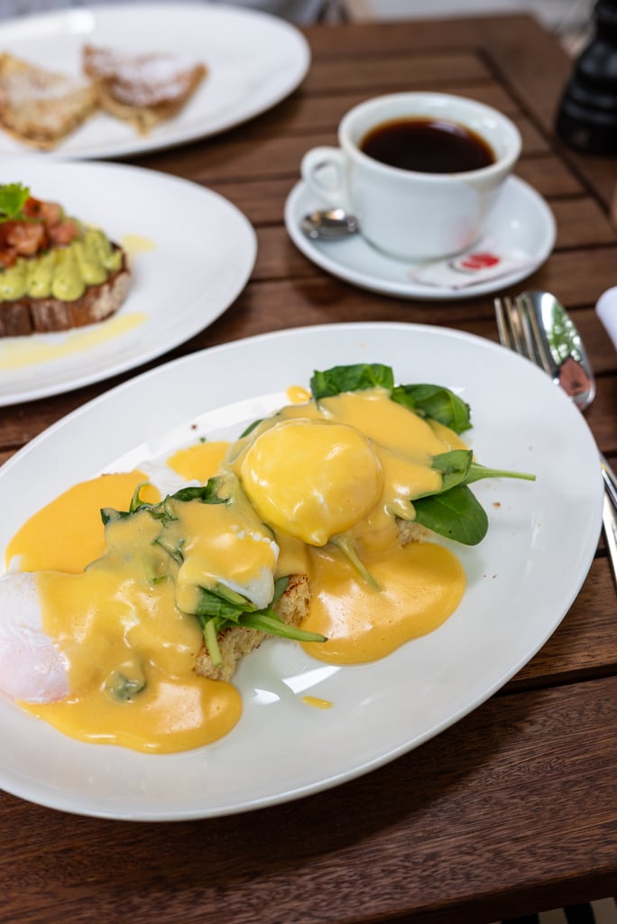 Plate of eggs Benedict with hollandaise and greens on a wooden table; a cup of coffee in the background.
