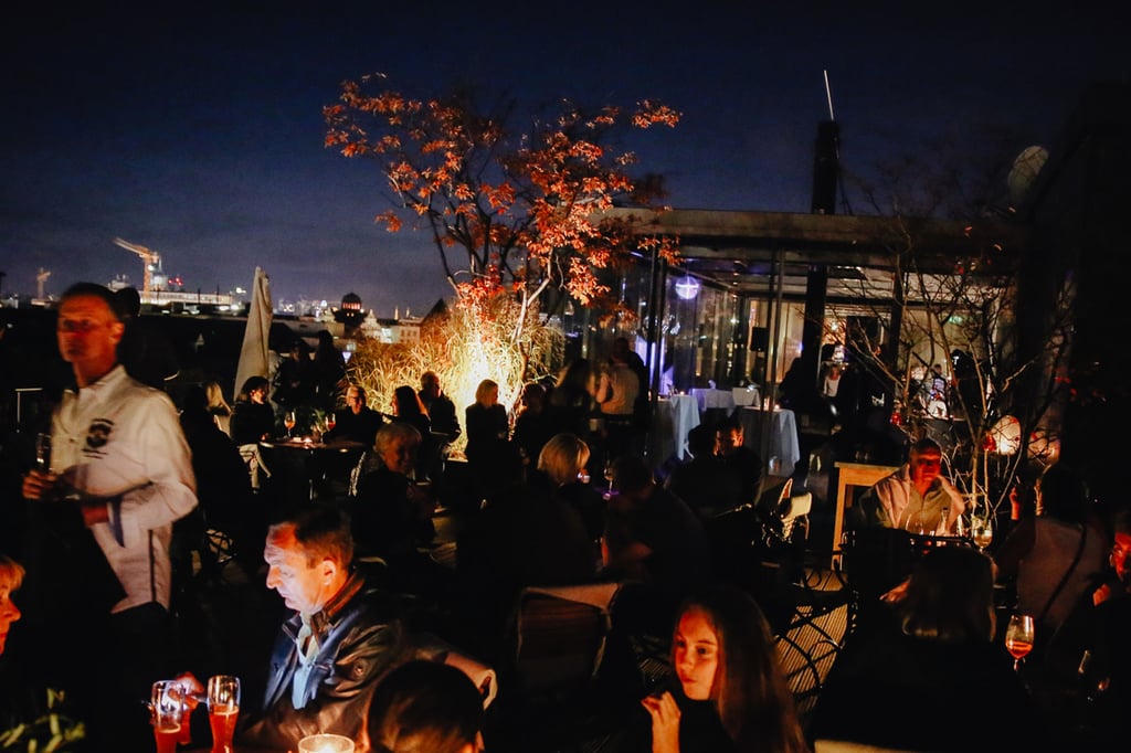 Nighttime rooftop restaurant scene with many diners, warm lighting, and a red-leafed tree against a dark sky.