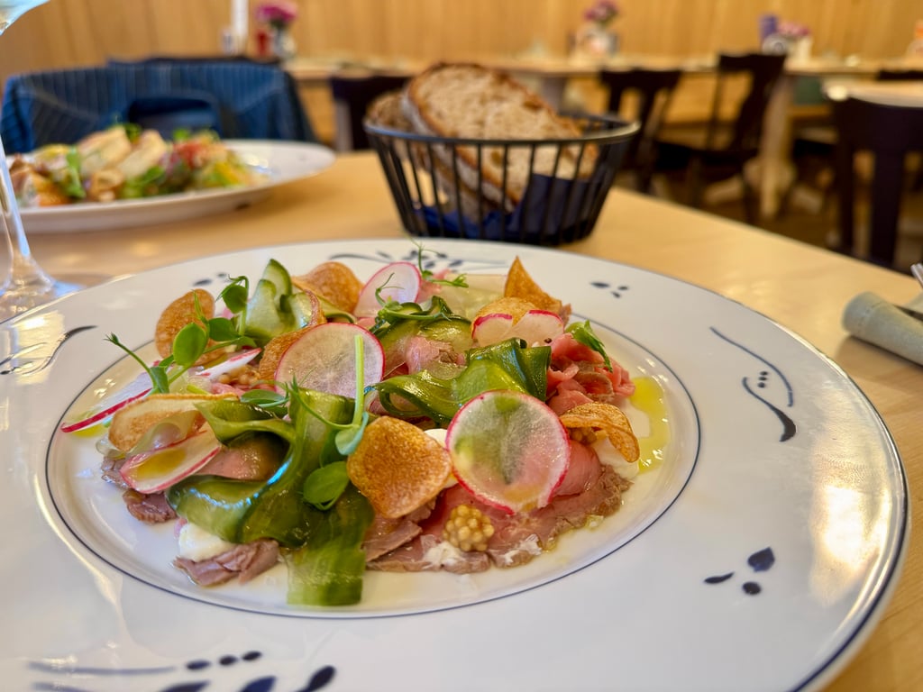 Plate of mixed greens salad with radish slices and crispy potato chips on a decorative white plate; a bread basket and dining room are blurred in the background.
